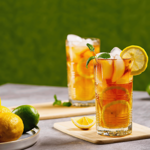 Two tall glasses of iced tea garnished with lemon slices, lime, mint, and ice, placed on wooden boards with fresh citrus fruit nearby against a green background.