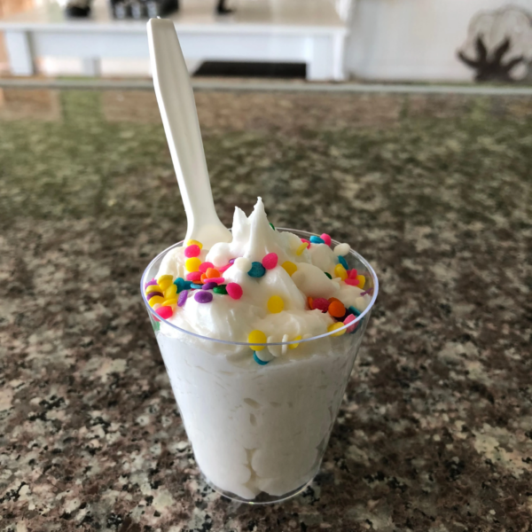A clear WNA Comet 1 oz portion cup filled with vanilla ice cream, topped with colorful sprinkles, and accompanied by a white plastic spoon, placed on a speckled countertop.