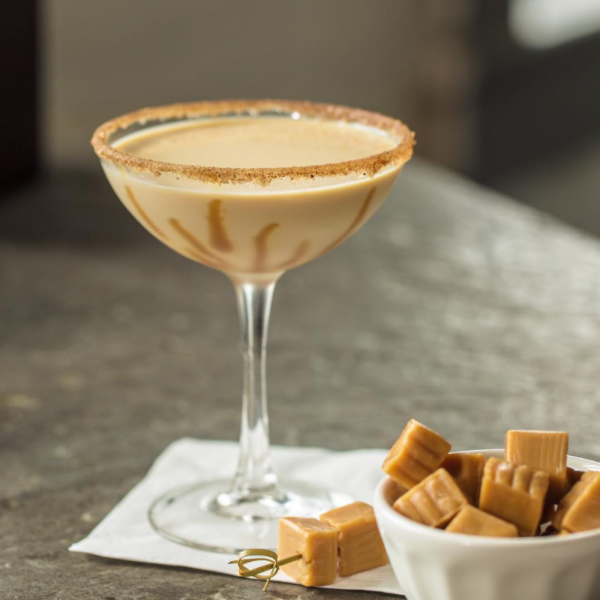 A cocktail glass with caramel drizzle and a sugared rim, accompanied by a small bowl of caramel candies, showcasing Monin Caramel Sauce as an ingredient.