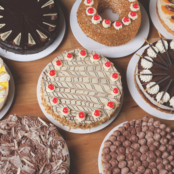 Image of multiple decorated cakes displayed on bright white cake circles. The cakes vary in flavors and designs, featuring chocolate, fruit, and cream toppings.