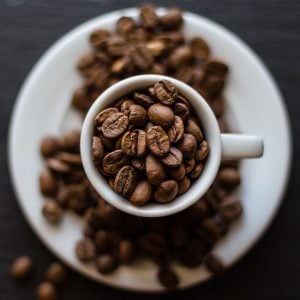 White coffee cup filled with whole coffee beans, placed on a matching white saucer also covered with coffee beans, set against a dark background.