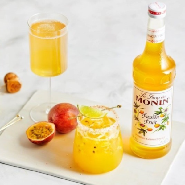 A bottle of Monin Passion Fruit Syrup next to two glasses filled with vibrant passion fruit drinks, garnished with lime, accompanied by fresh passion fruit halves on a marble surface.
