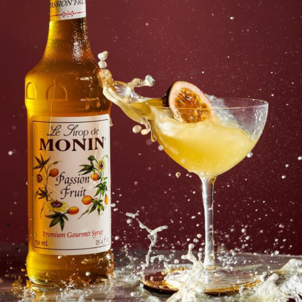A bottle of Monin Passion Fruit Syrup beside a cocktail glass filled with a passion fruit drink, garnished with a passion fruit slice, with a dramatic splash effect against a deep red background.